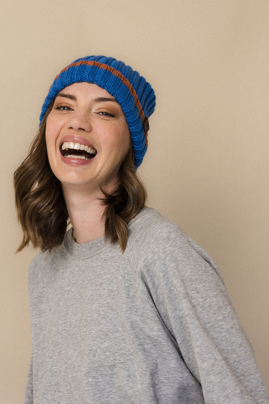 A person wearing a striped merino wool beanie in blue and cinnamon colors, smiling against a beige background.