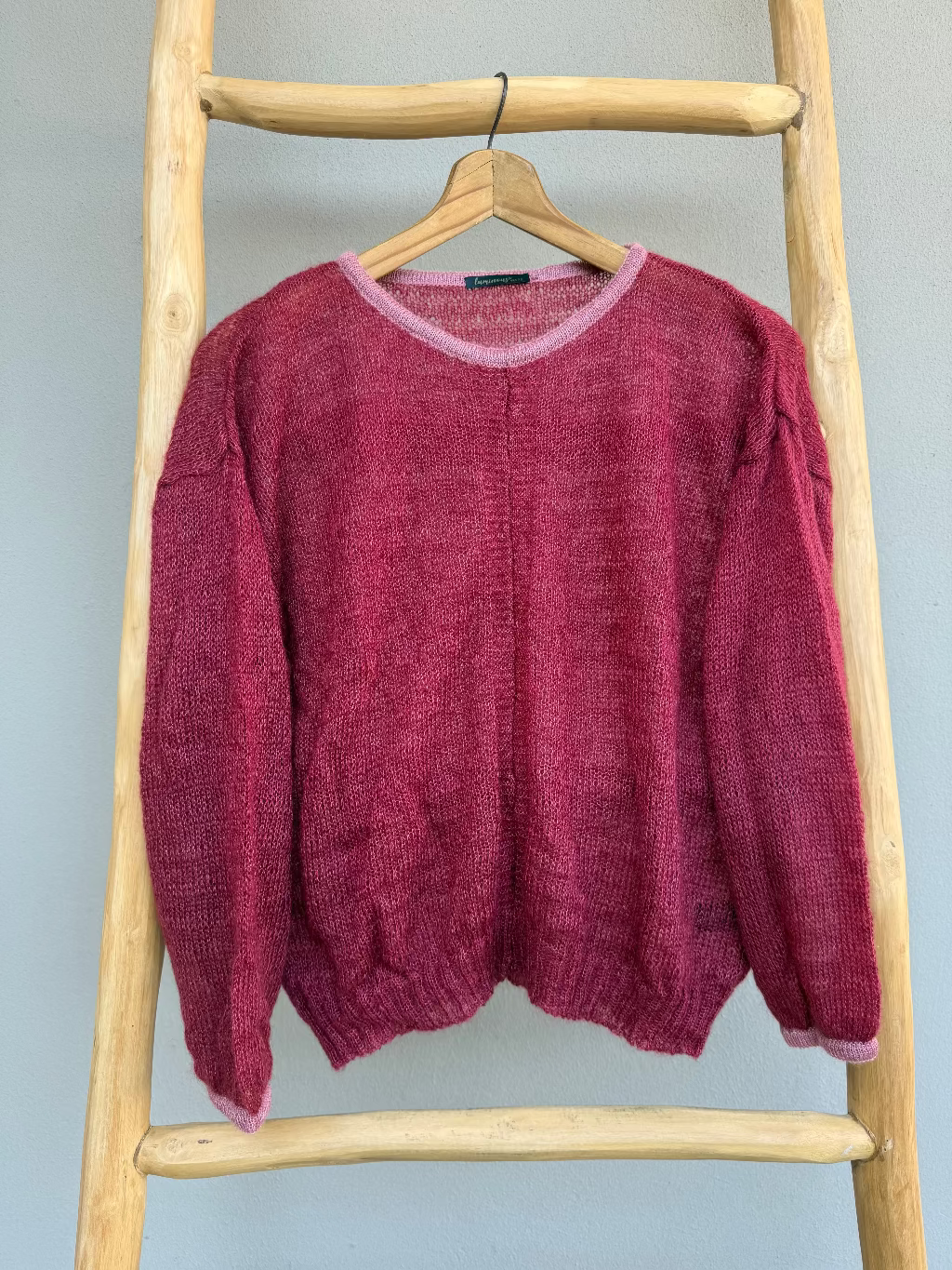 A textured mohair knit sweater in a cherry colour with dusty pink trims, hanging on a wooden hanger from a rustic wooden ladder against a light gray wall.