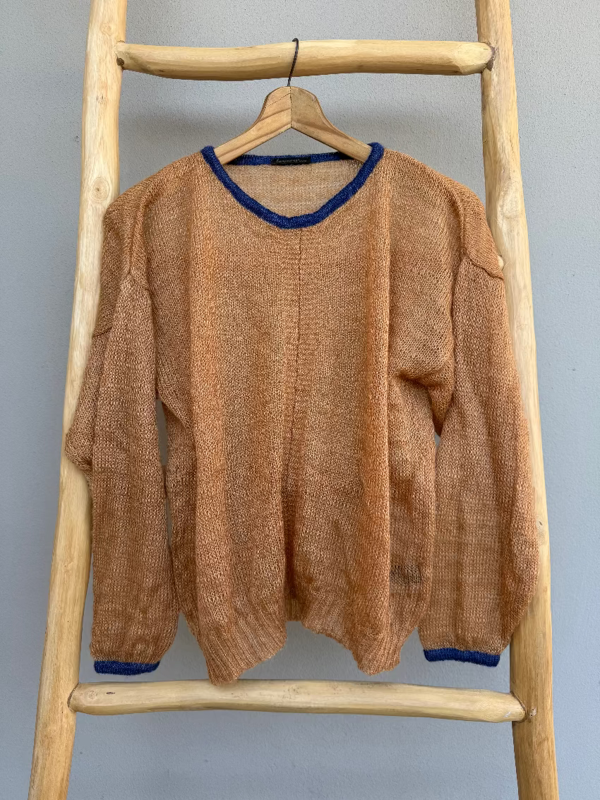 A cinnamon textured knit mohair sweater with navy trim on the collar and cuffs, hanging on a wooden hanger from a rustic wooden ladder against a gray wall.