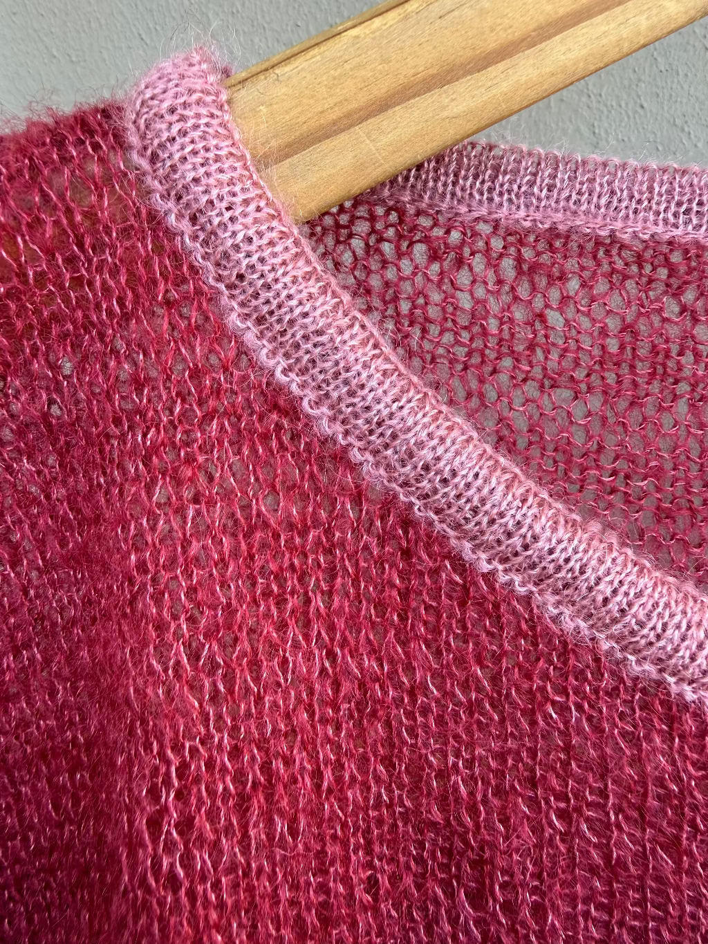Close-up of a mohair open-knit sweater in a cherry tone, with a contrasting light pink ribbed collar, hanging on a wooden hanger against a gray wall.