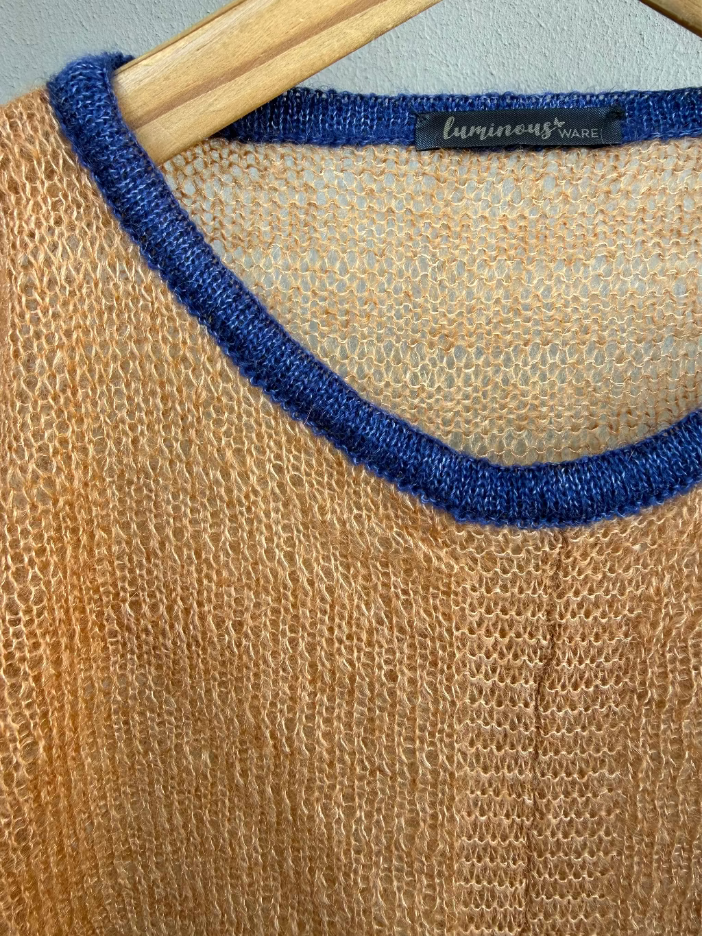 A close-up of a cinnamon knit mohair sweater with a contrasting dark blue ribbed crew neck, hanging on a wooden hanger. The brand label 'luminous ware' is visible at the collar.