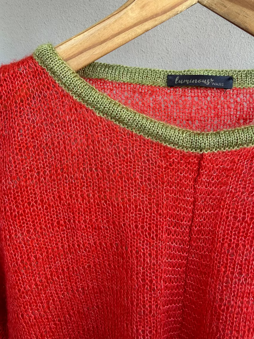 A close-up of a tangerine knit sweater hanging on a wooden hanger against a light gray wall. The sweater has an olive green neck and a black brand label reading 'luminous ware' visible inside the collar.