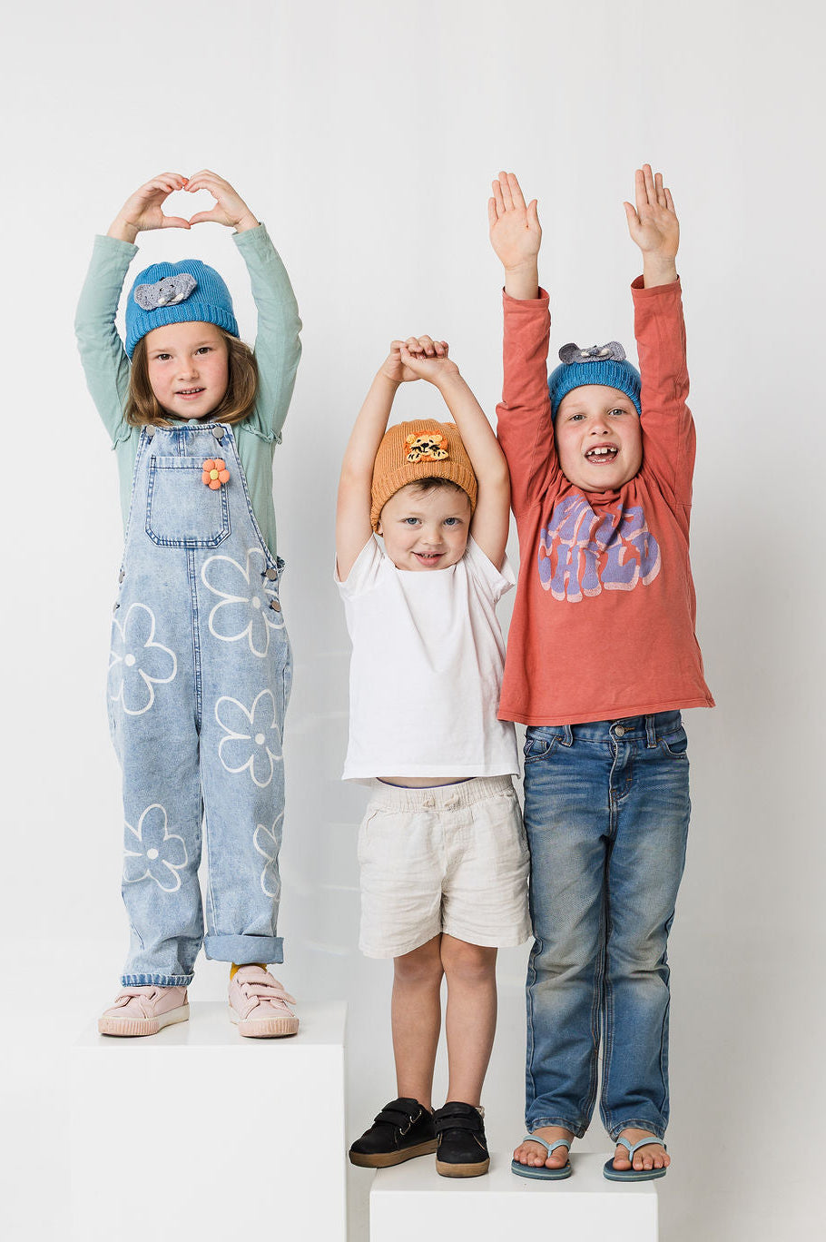 Three children standing on white blocks against a white background, wearing colorful clothing and animal beanies.