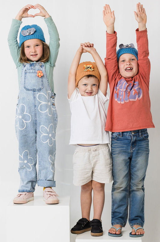 Three children standing on white blocks against a white background, wearing colorful clothing and animal beanies.