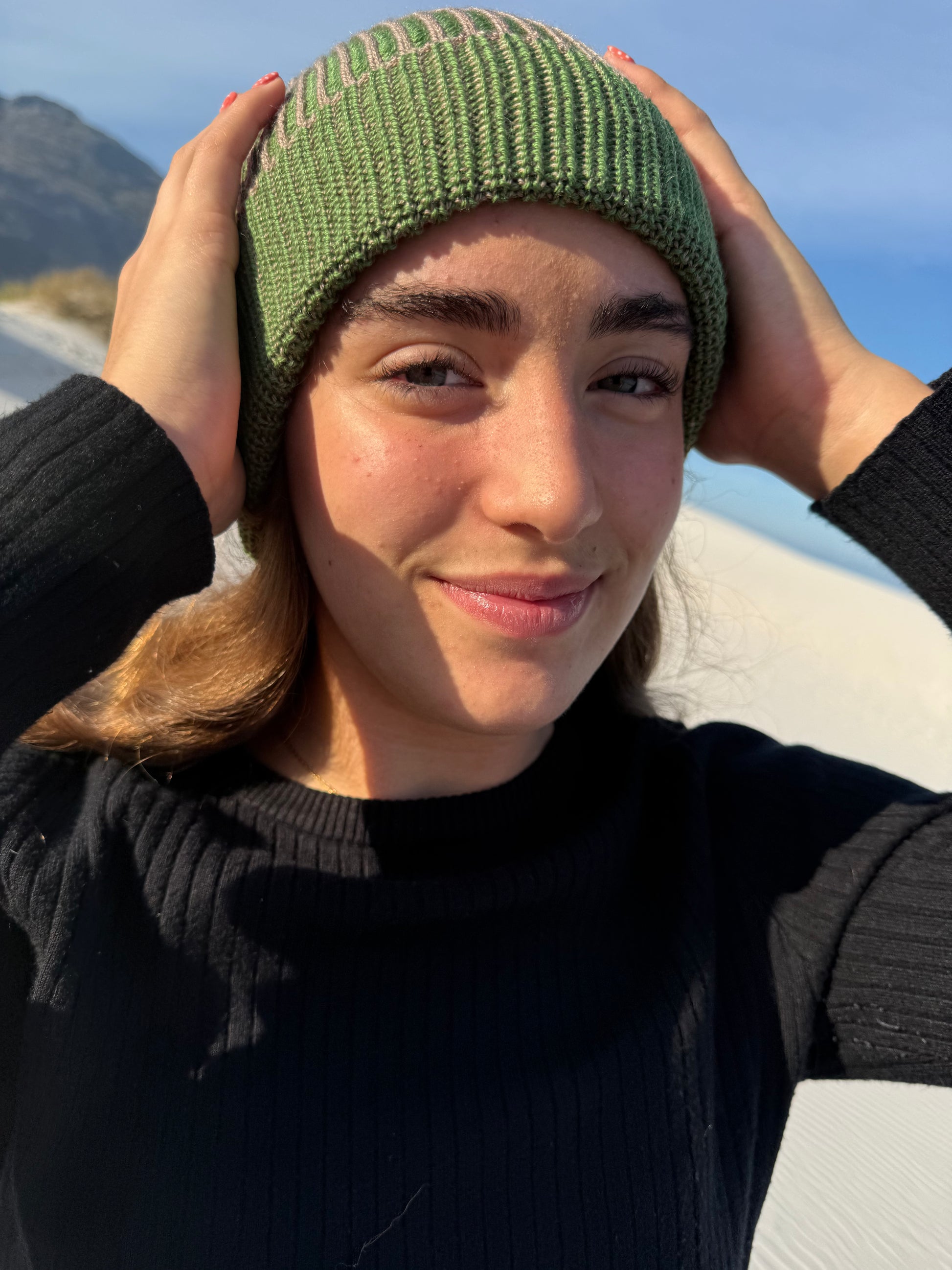 A woman wearing a brioche knit green beanie hat and a black ribbed sweater, posing outdoors with her hands on her head against a sunny beach background.