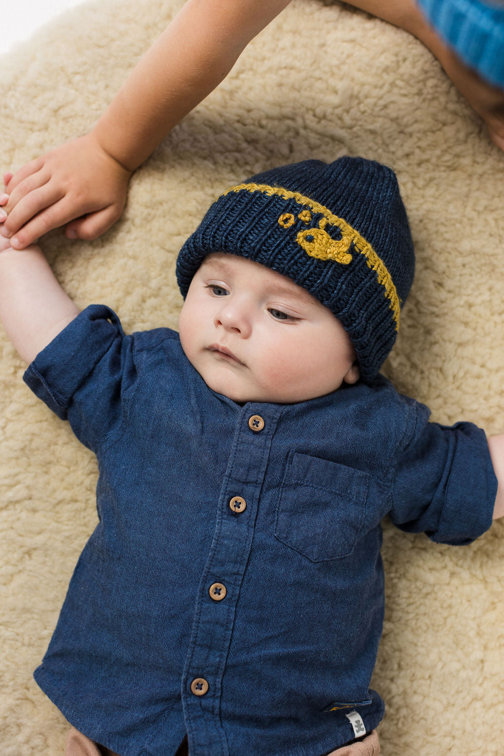 Baby wearing a blue wool beanie with crochet fish detail and matching outfit lying on a beige rug.