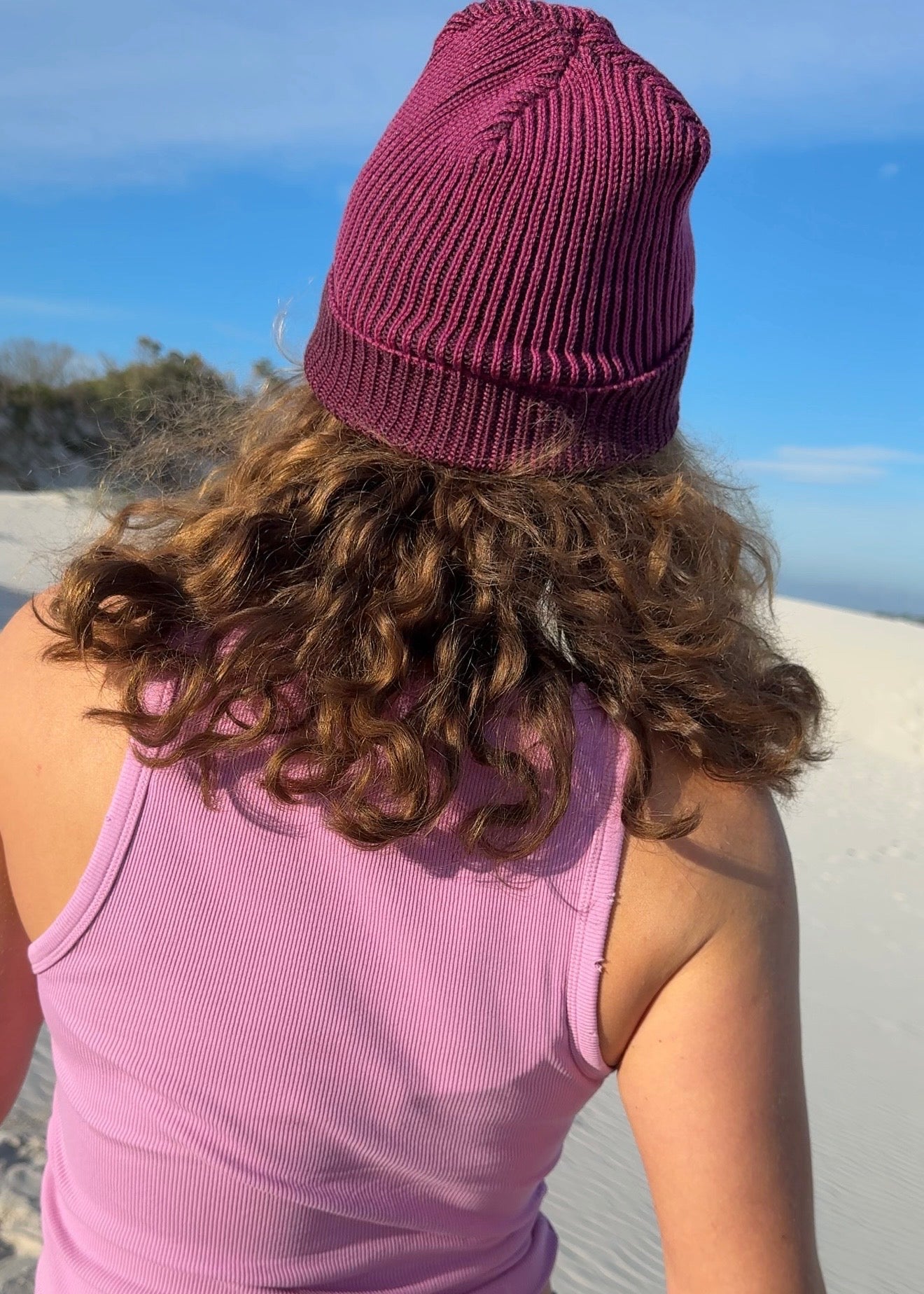A person viewed from behind wearing a brioche knit plum beanie, a lavender ribbed tank top, standing outdoors on a sandy dune under a blue sky.