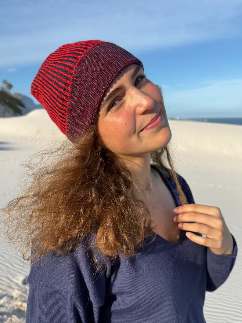 A woman with long wavy brown hair wearing a red and navy brioche knit beanie and a navy blue top, standing outdoors on a sandy dune under a blue sky.